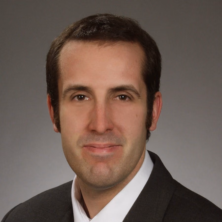 Headshot of a man with short dark hair in a suit jacket and white shirt against a gray background.
