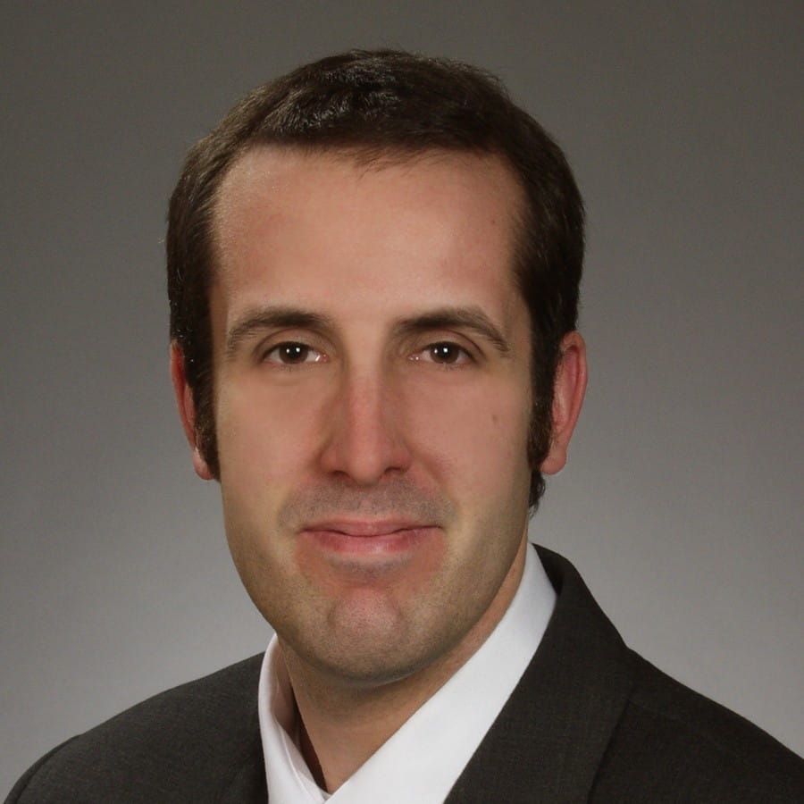 Headshot of a man with short dark hair in a suit jacket and white shirt against a gray background.