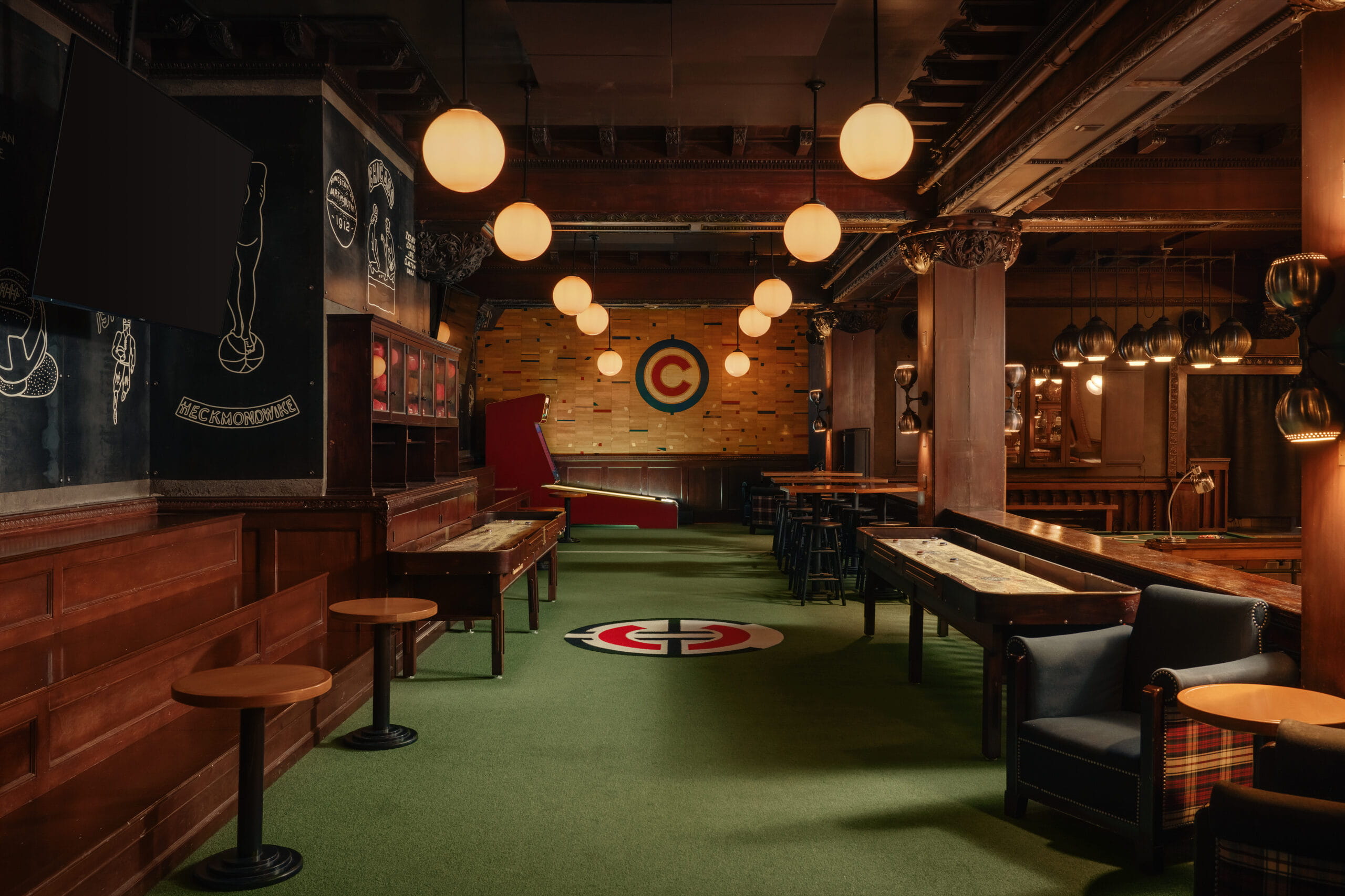 Cozy game room with shuffleboard tables, arcade machine, round hanging lights, and wood-paneled walls.