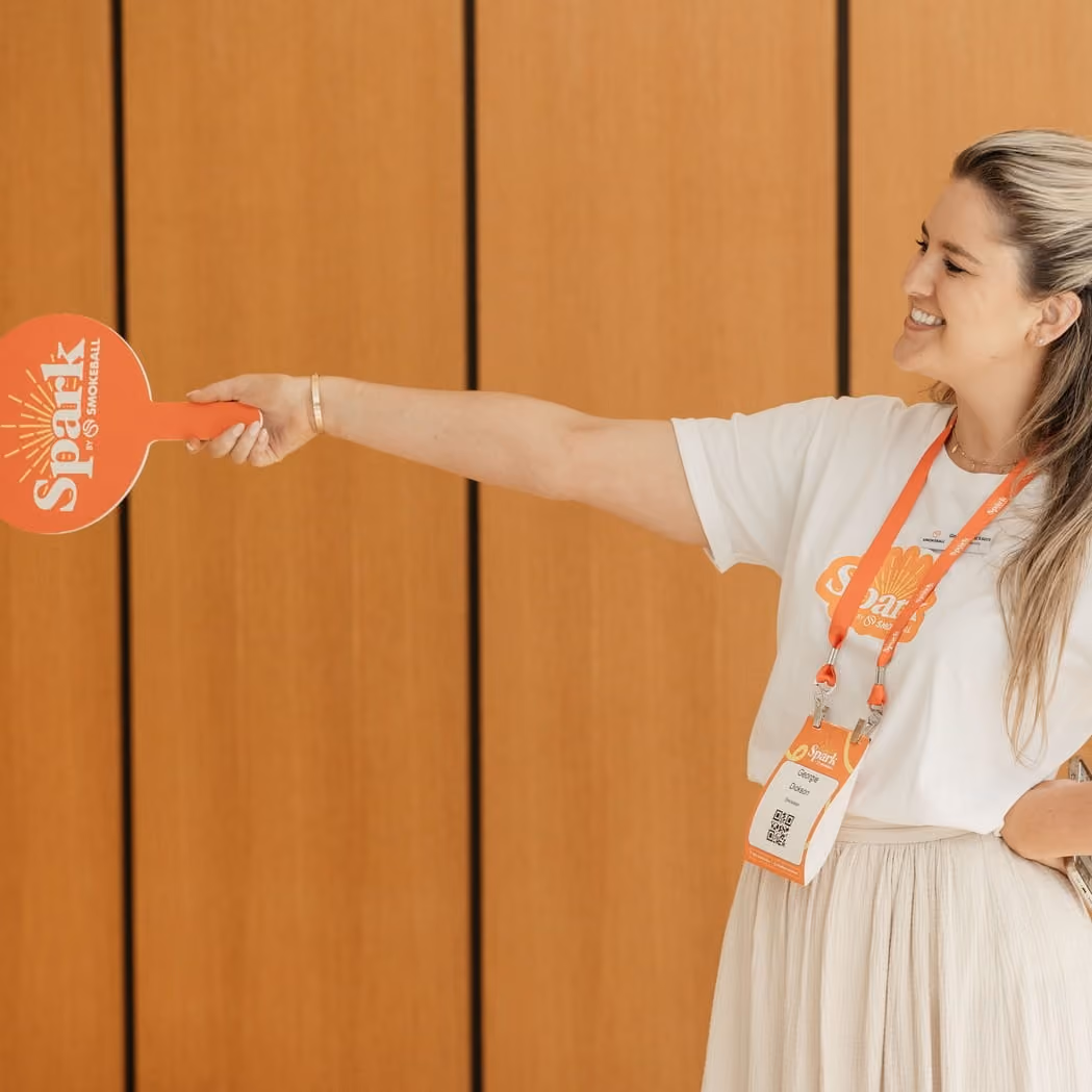 Smiling woman holding an orange 'Spark by Smokeball' paddle, wearing a matching lanyard and badge.