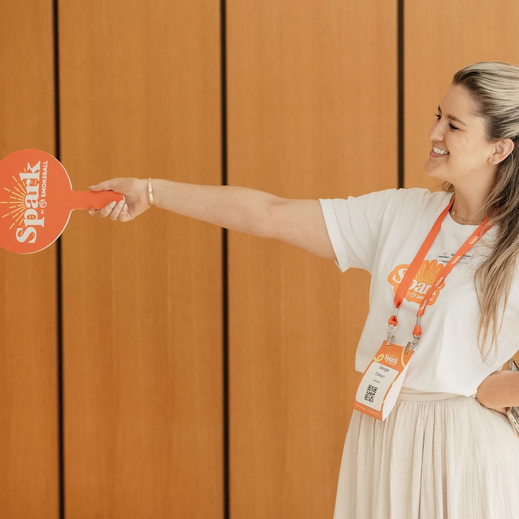 Smiling woman holding an orange 'Spark by Smokeball' paddle, wearing a matching lanyard and badge.
