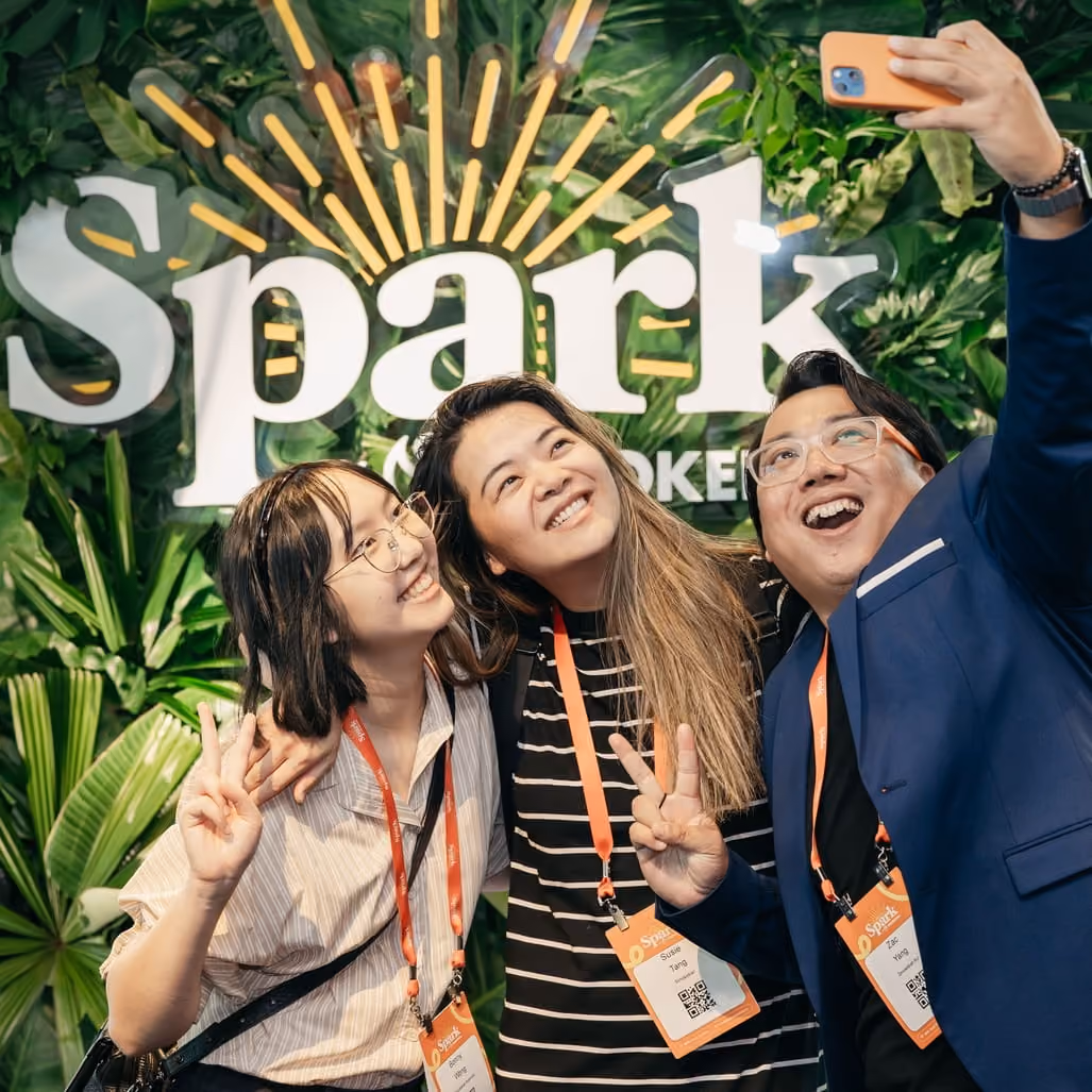 Three people smiling and posing for a selfie with peace signs in front of a green leafy Spark backdrop.