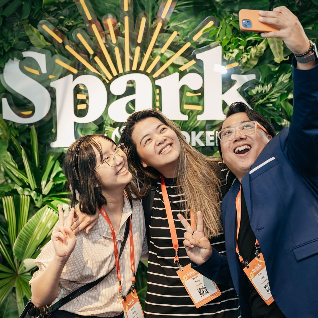 Three people smiling and posing for a selfie with peace signs in front of a green leafy Spark backdrop.