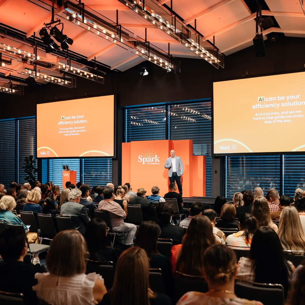 Man in a light jacket presenting on stage with orange backdrop and two screens about AI efficiency solution.
