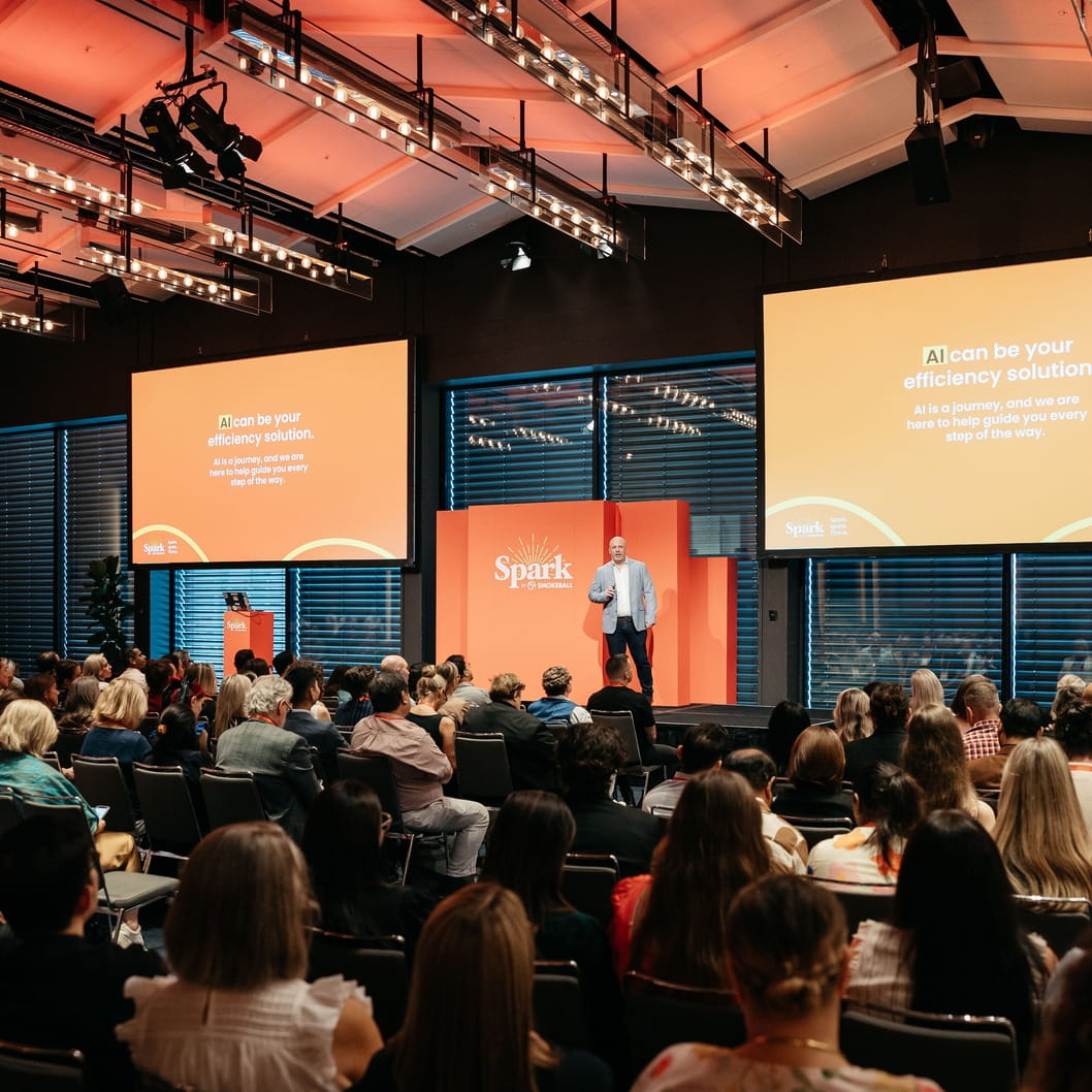 Man in a light jacket presenting on stage with orange backdrop and two screens about AI efficiency solution.
