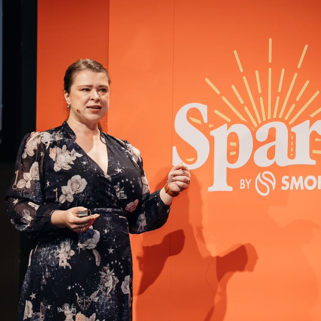 A woman in a black floral dress speaking with a microphone against an orange background with logo text.
