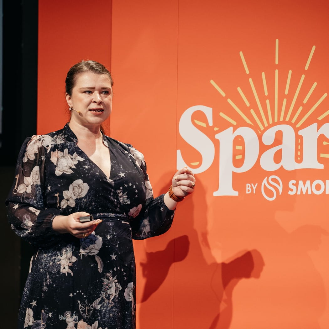 A woman in a black floral dress speaking with a microphone against an orange background with logo text.