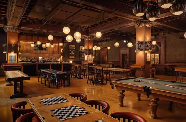 Warmly lit bar with pool table, shuffleboard, checker tables, and hanging globe lights.