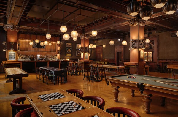 Warmly lit bar with pool table, shuffleboard, checker tables, and hanging globe lights.
