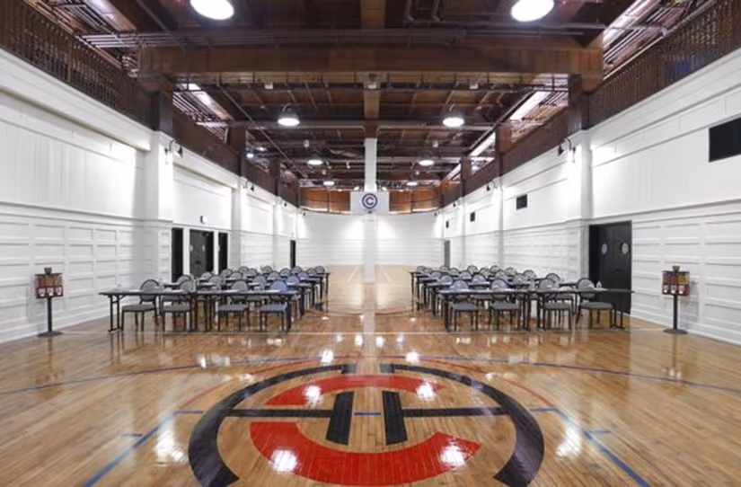 Indoor basketball court with hardwood floor, tables and chairs set up along both sides under a wooden ceiling.