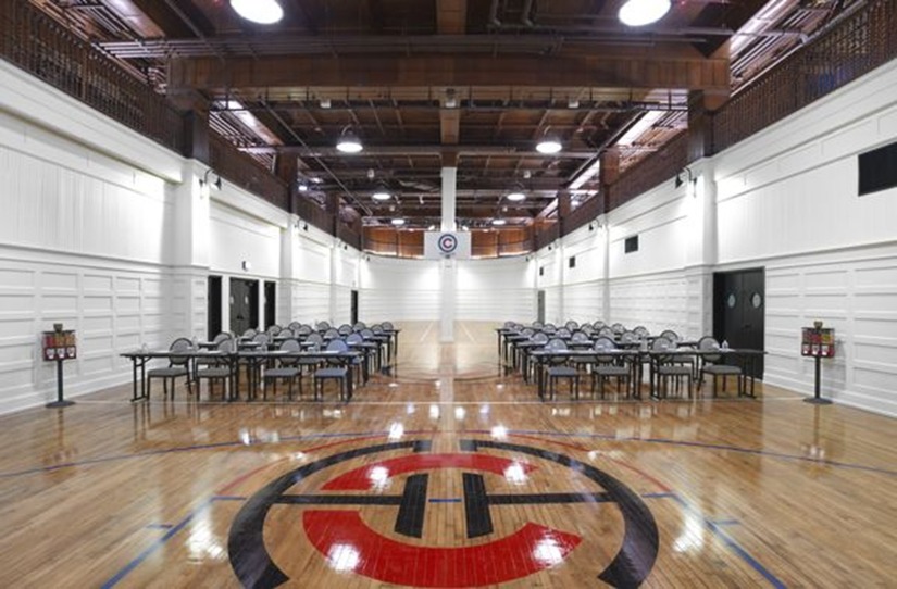 Indoor basketball court with hardwood floor, tables and chairs set up along both sides under a wooden ceiling.