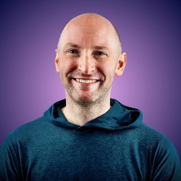 Smiling bald man wearing a teal hoodie with a purple gradient background.