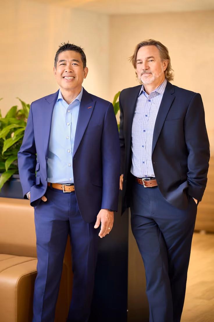 Two men in business suits standing indoors with relaxed expressions near a plant and sofa.