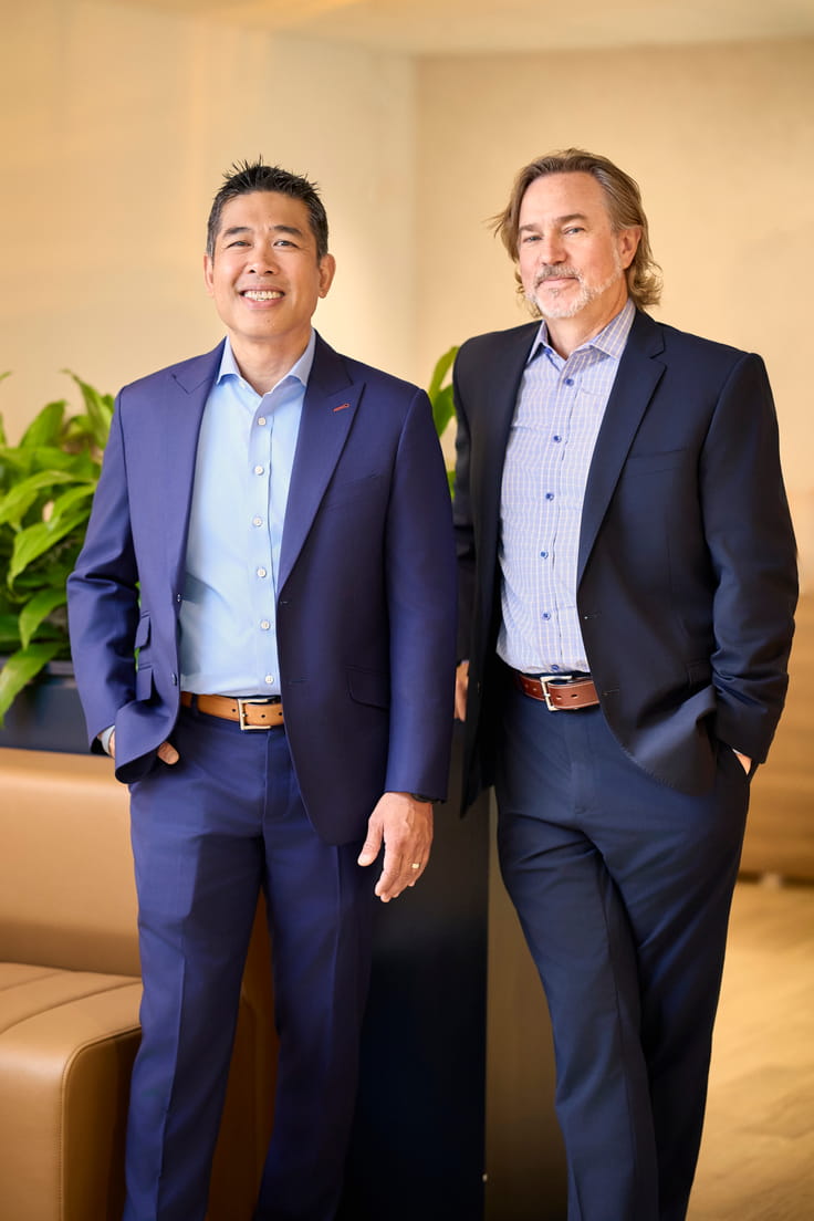Two men in business suits standing indoors with relaxed expressions near a plant and sofa.