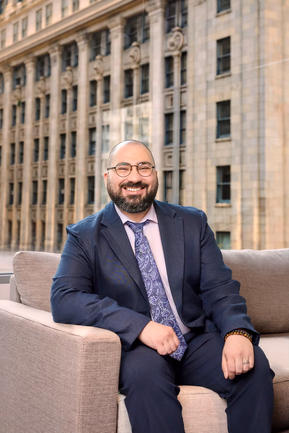 Smiling man with glasses and beard wearing a suit and tie sitting on a beige couch by a window.
