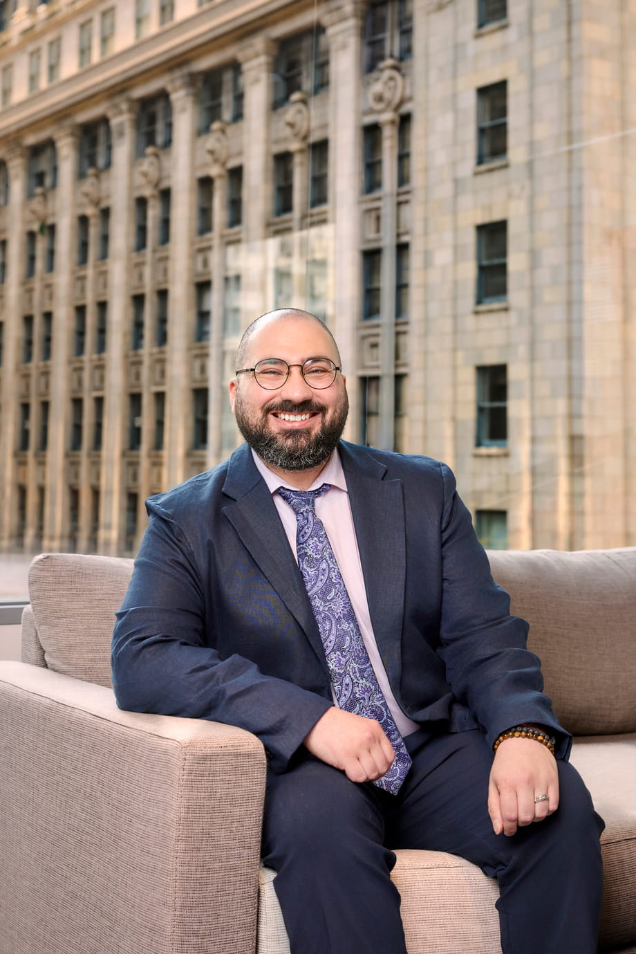 Smiling man with glasses and beard wearing a suit and tie sitting on a beige couch by a window.