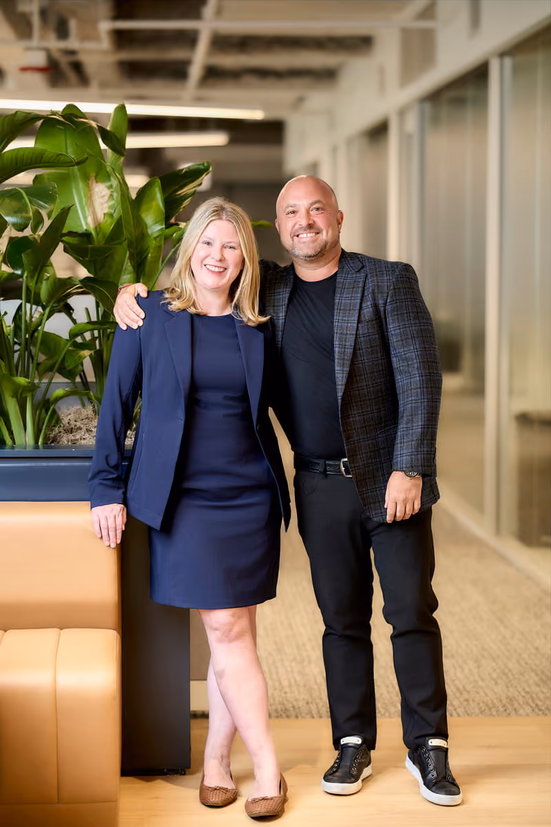 Smiling man and woman stand side by side in office corridor with plants and leather seating.