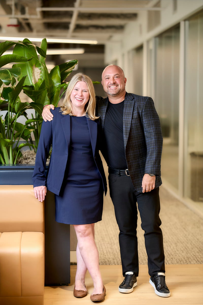 Smiling man and woman stand side by side in office corridor with plants and leather seating.
