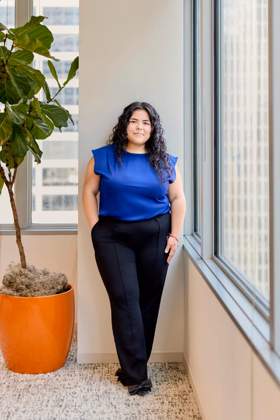 Woman in blue blouse and black pants standing near a window with a large potted plant nearby.