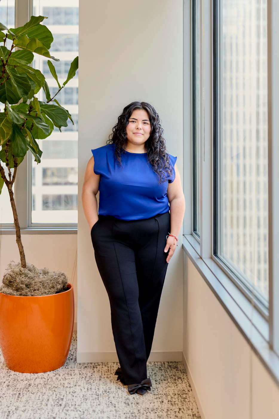 Woman in blue blouse and black pants standing near a window with a large potted plant nearby.