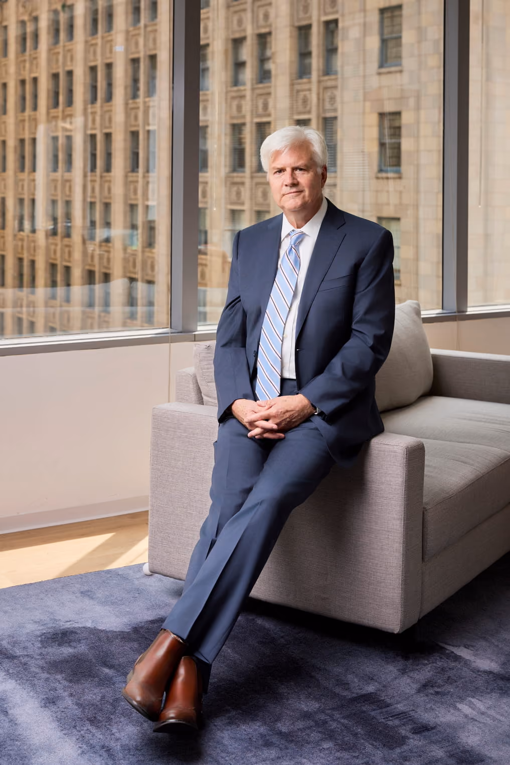 Older man in a blue suit and striped tie sitting on the arm of a gray couch in an office with city views.