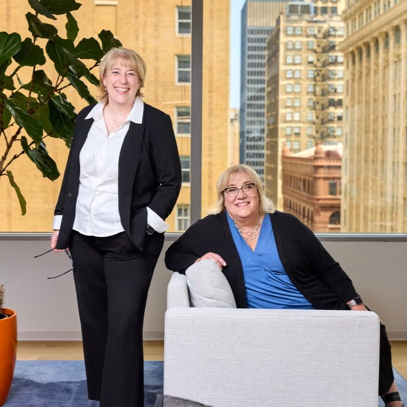 Two women smiling in a bright office with city buildings visible through a window behind them.