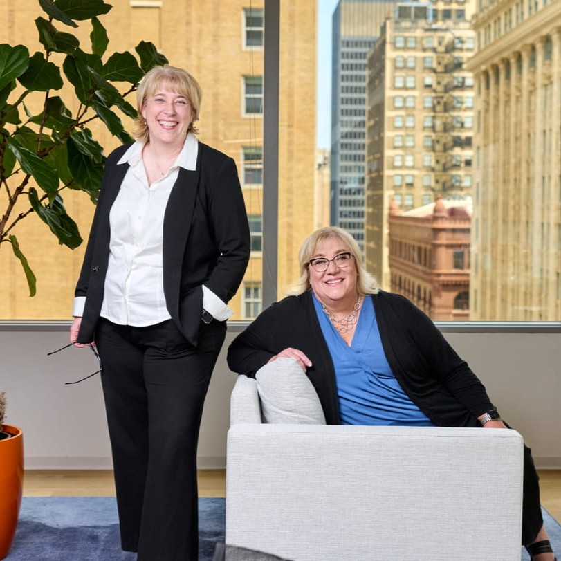 Two women smiling in a bright office with city buildings visible through a window behind them.