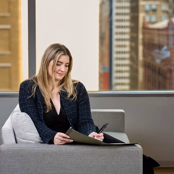Woman in dark jacket sitting on gray sofa writing on a clipboard with city buildings in the background.