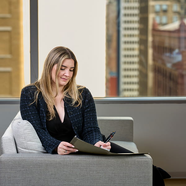Woman in dark jacket sitting on gray sofa writing on a clipboard with city buildings in the background.