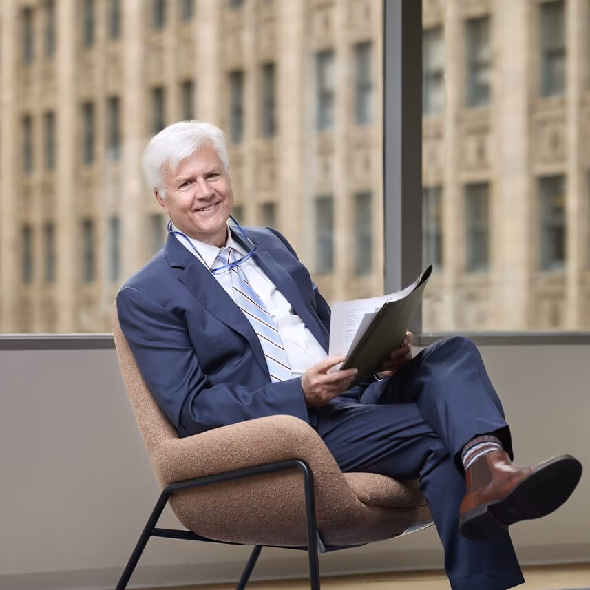 Smiling older man with white hair in a blue suit seated, holding papers by a large window.