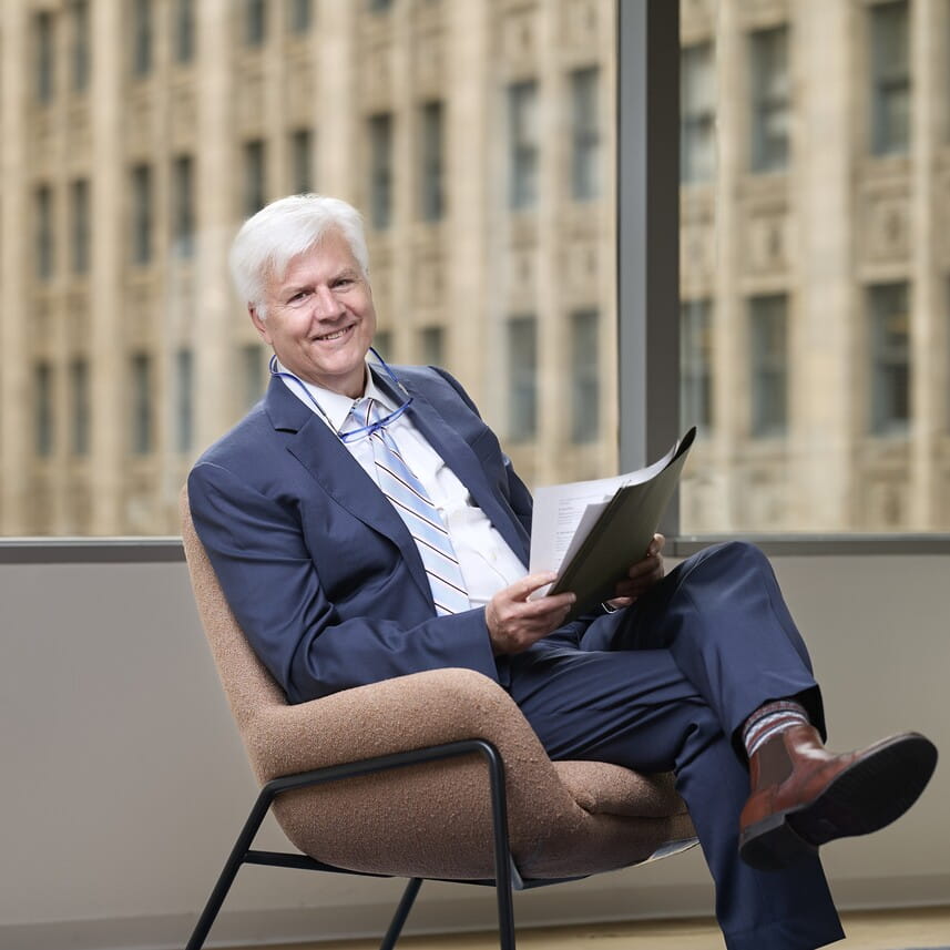 Smiling older man with white hair in a blue suit seated, holding papers by a large window.