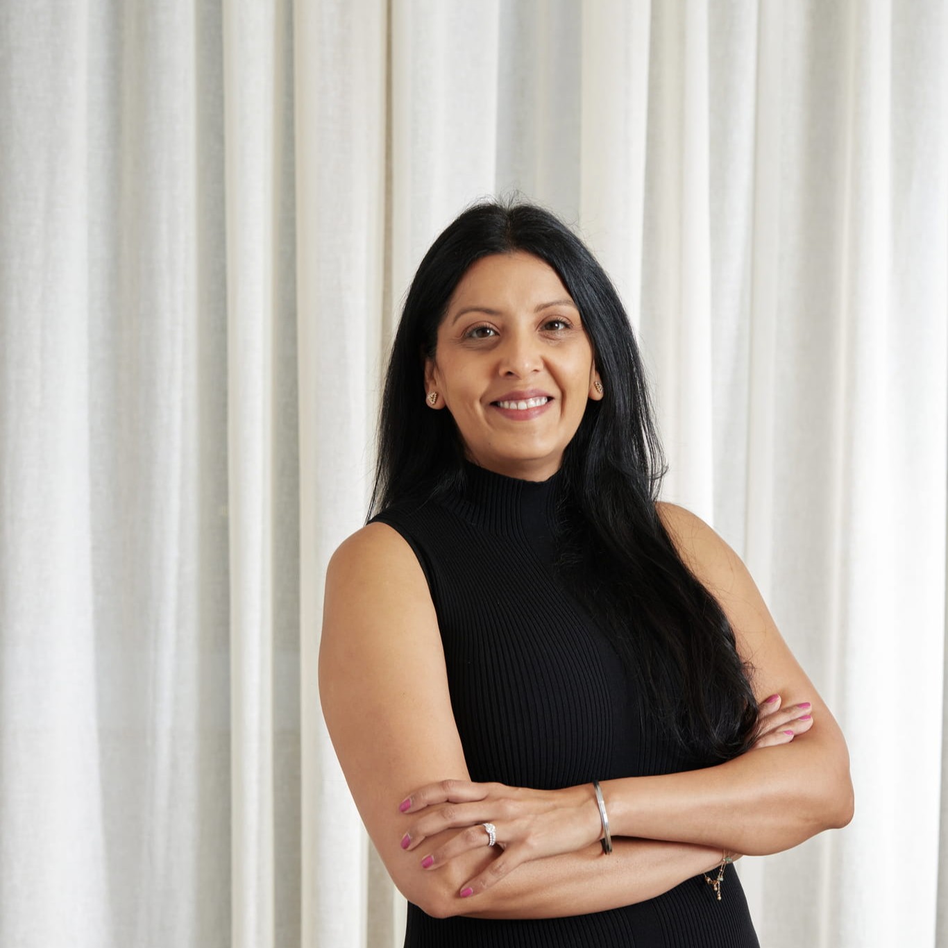 Smiling woman with long black hair in a sleeveless black top, arms crossed, standing by white curtains.