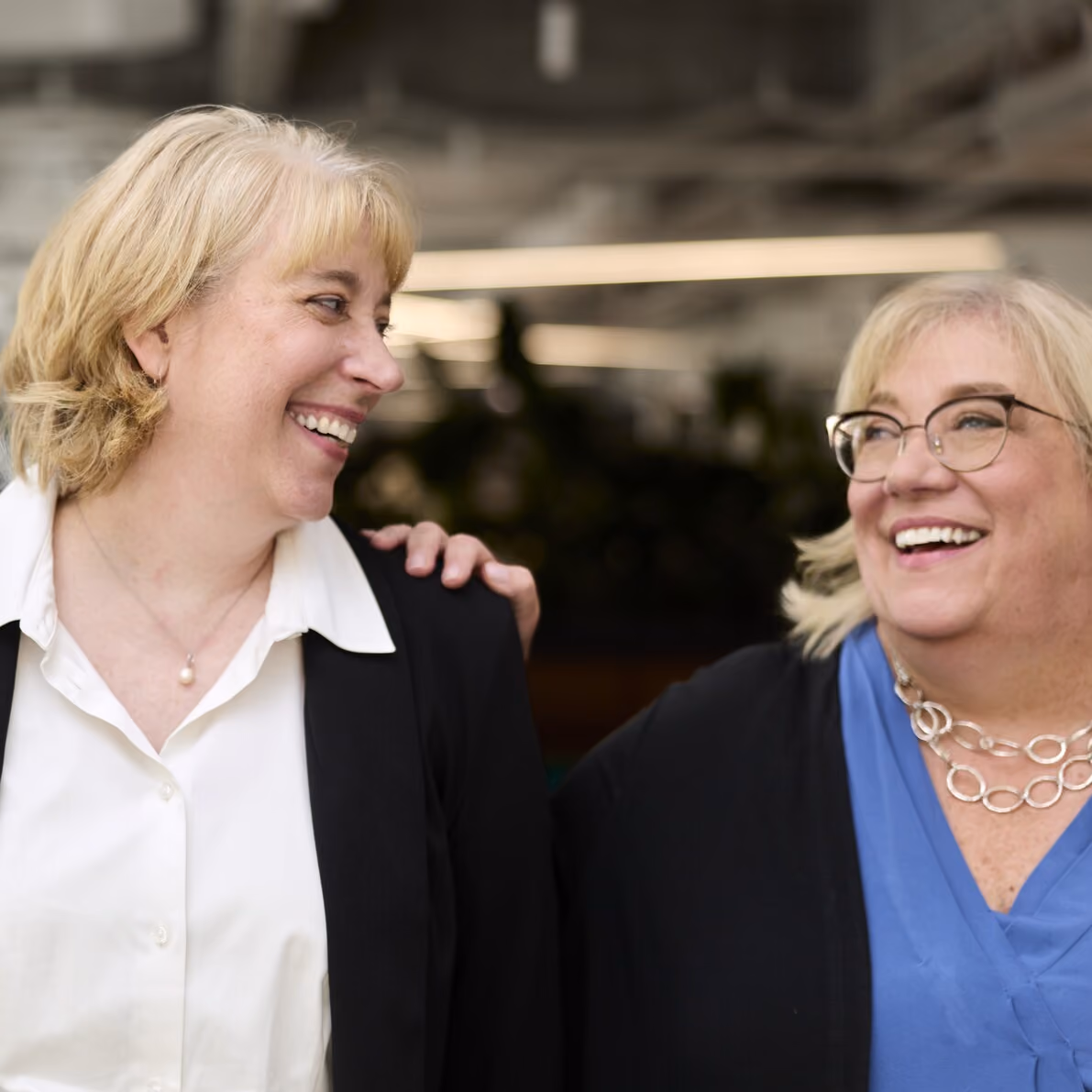 Two middle-aged women smiling and looking at each other in a friendly indoor setting.