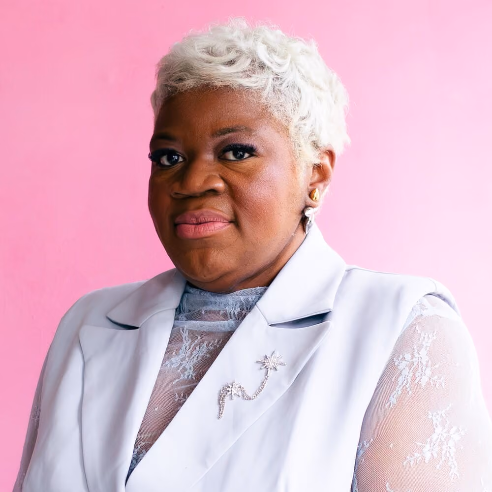 Woman with short curly white hair wearing white lace and vest against a pink background.