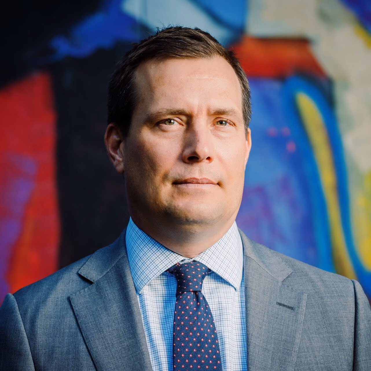 Serious man in gray suit and checkered shirt with patterned tie standing before colorful abstract wall.