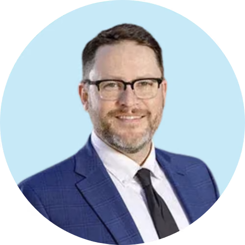 Man with glasses and beard wearing a blue suit and black tie smiling on a light blue background.