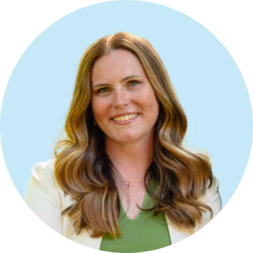 Smiling woman with long wavy brown hair wearing green top and white blazer.