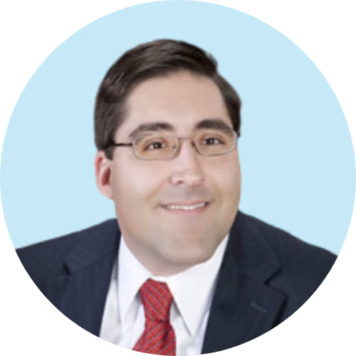 Man with glasses wearing a dark suit, white shirt, and red tie smiles against a light blue background.