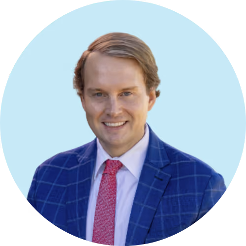 Smiling man with light brown hair wearing a blue checkered suit and red tie on light blue background.