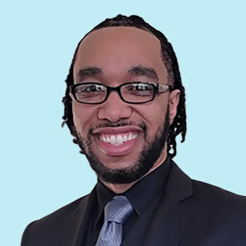 Smiling man with glasses wearing a black suit, black shirt, and gray tie against a light blue background.