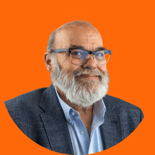 Smiling older man with white beard and glasses wearing a blazer and light blue shirt on orange background.