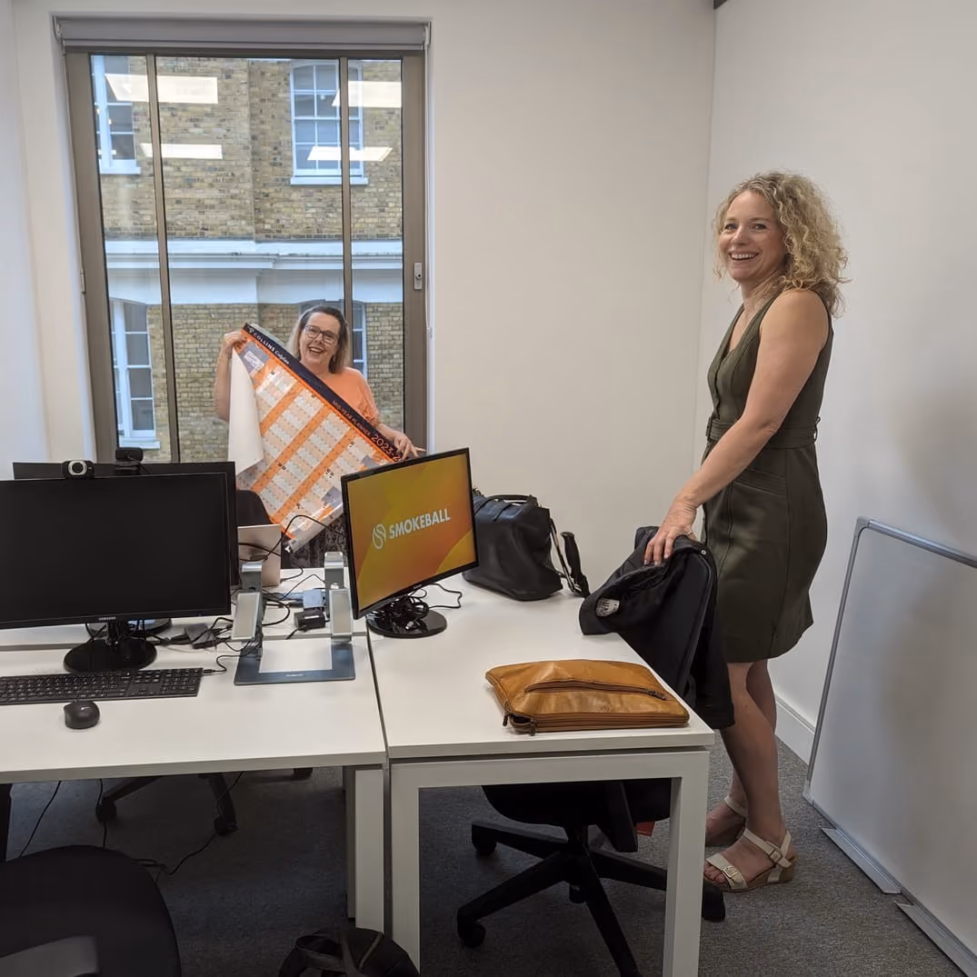 Two women in a modern office, one smiling by a desk and the other showing a large calendar.