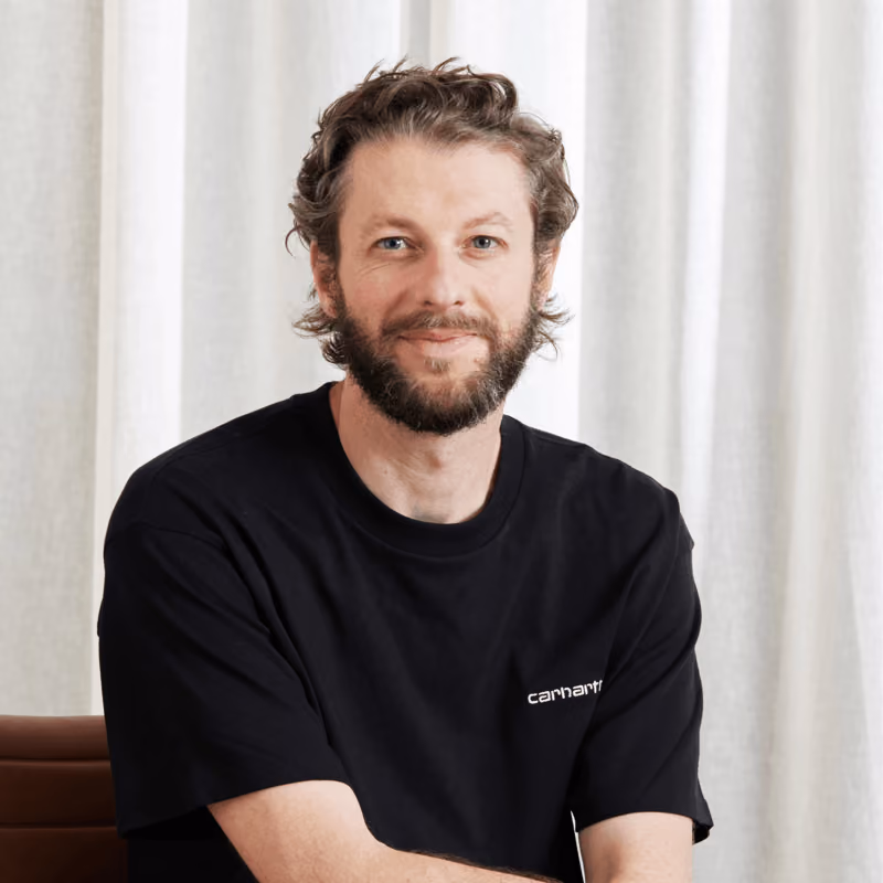 Man with beard and curly hair wearing a black Carhartt t-shirt sitting against white curtains.