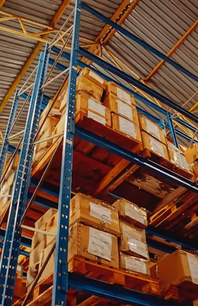 High-bay racking in a warehouse with several pallets full of stacked brown boxes, beneath a vaulted ceiling
