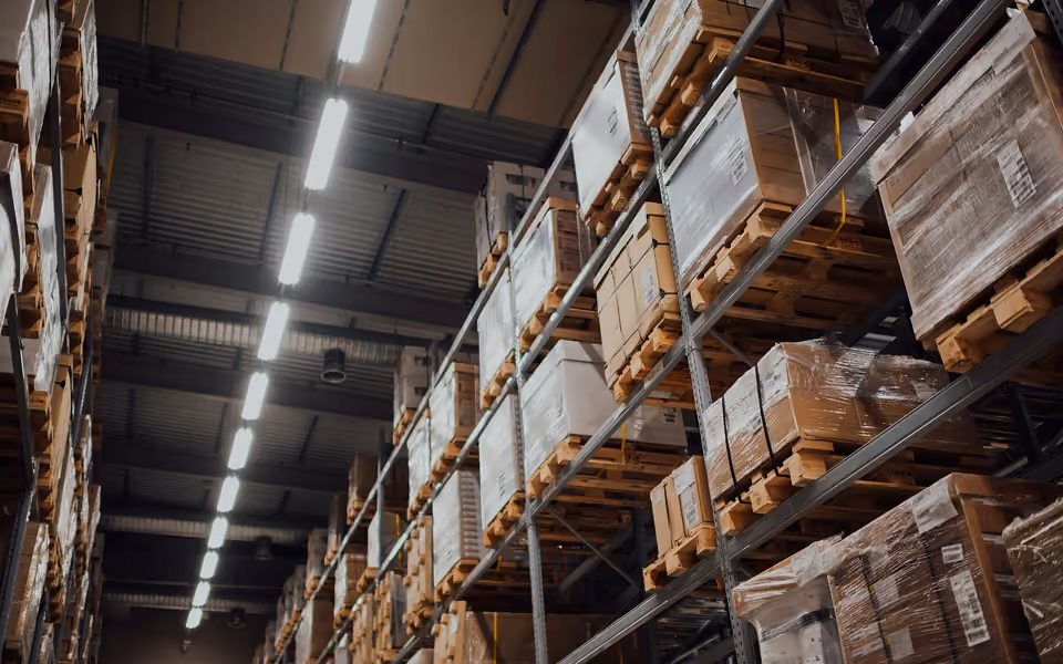 View into a warehouse with high racking filled with various boxes and pallets, lit by rows of ceiling lights