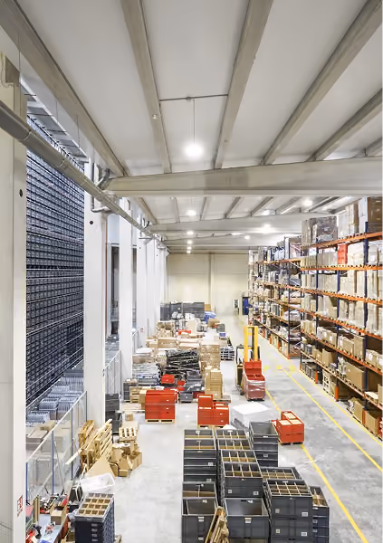 Large, well-lit warehouse with high racking full of boxes on the right and many pallets and crates on the floor