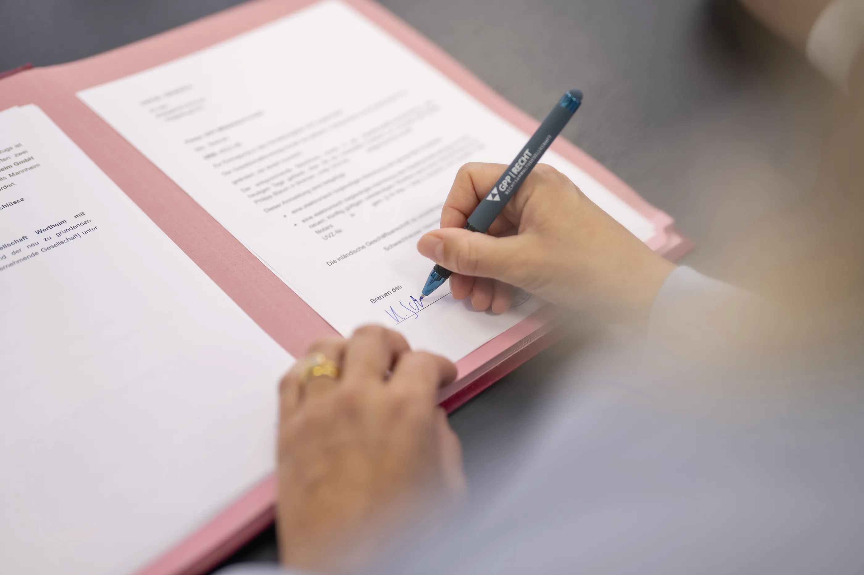 Person unterschreibt ein Dokument mit einem blauen Stift auf einem rosafarbenen Ordner.