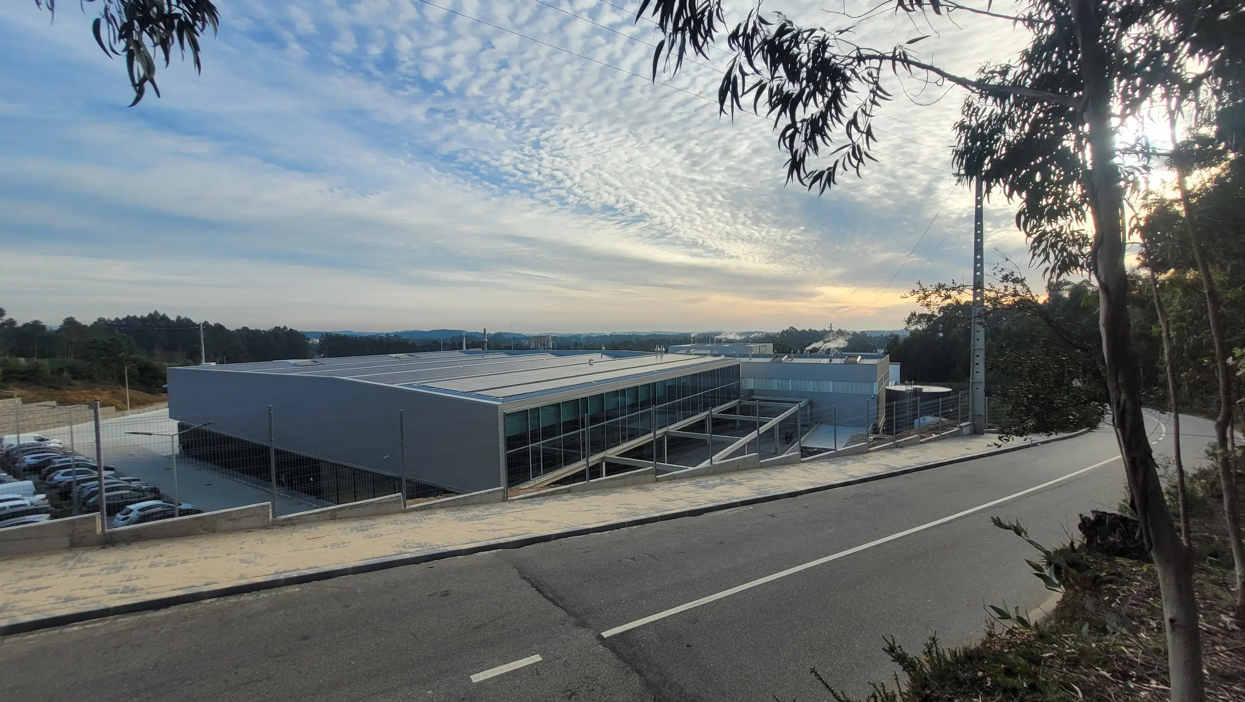 Modern industrial building with large glass windows, surrounded by a fence, a paved road curving around on the right, under a partly cloudy sky during sunset.
