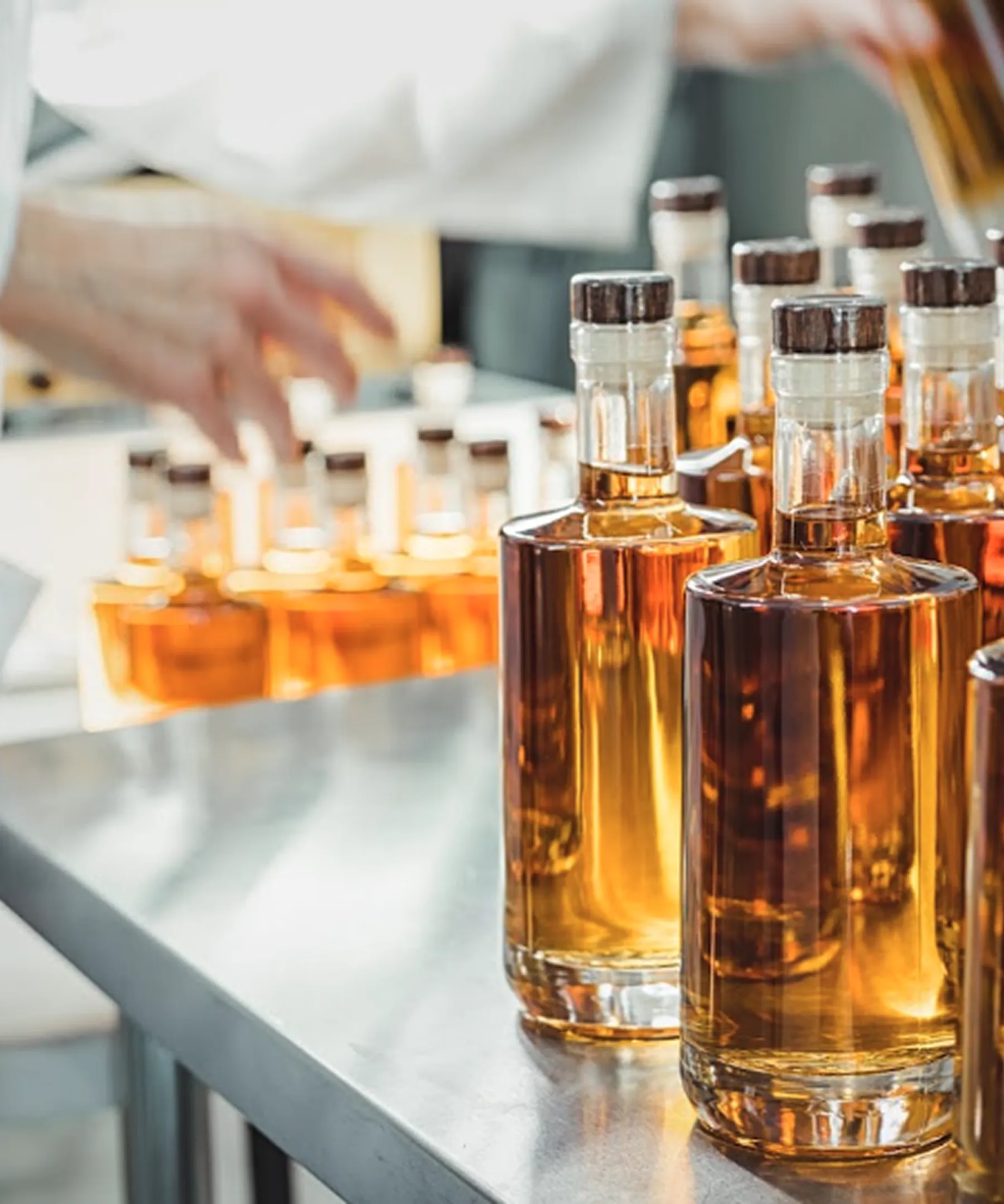 Close-up of multiple glass bottles arranged on a metal table with a blurred person handling more bottles in the background.