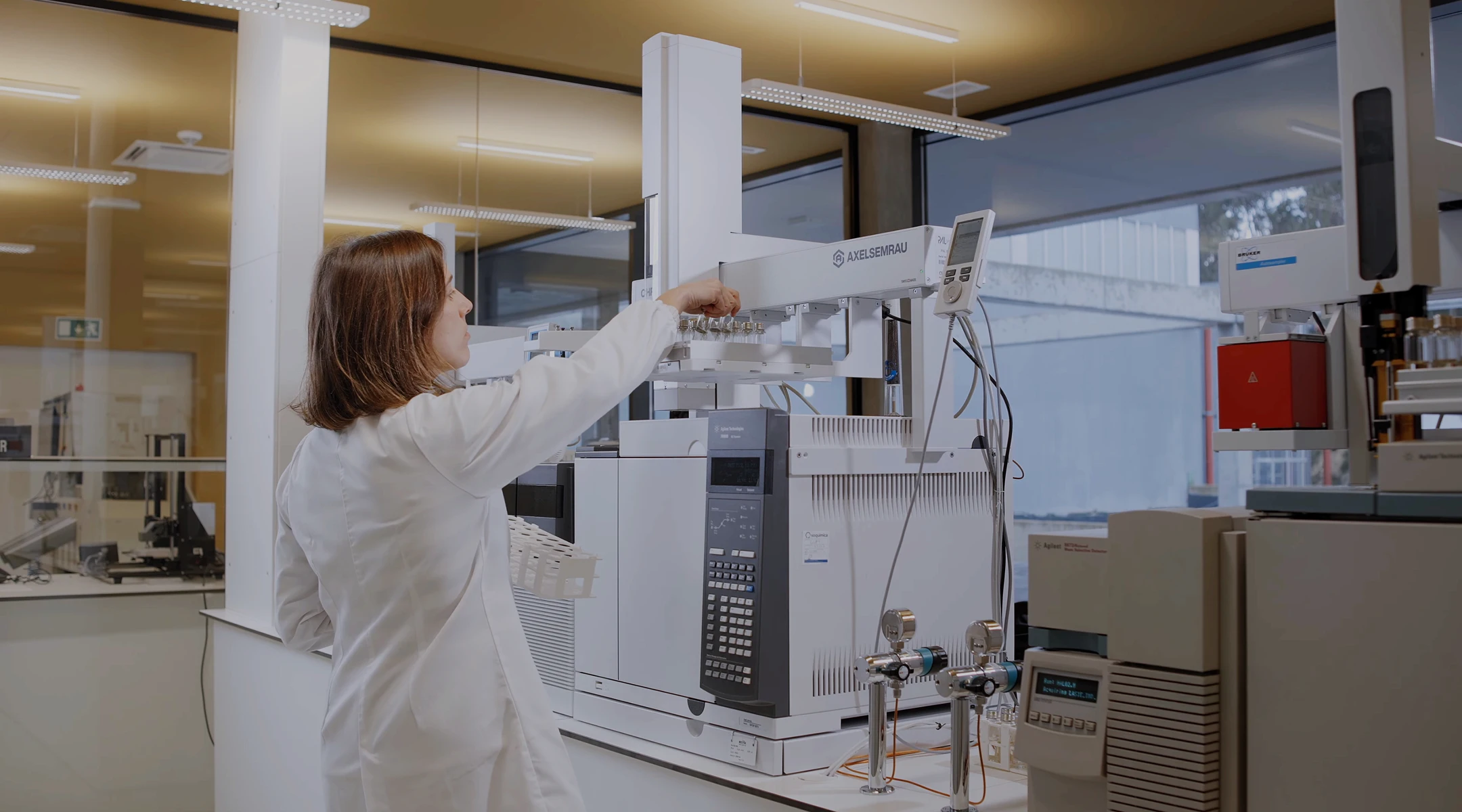A woman in a white lab coat operates a large laboratory analyzer machine labeled AXELSEMBAU in a modern lab with scientific equipment and large windows.